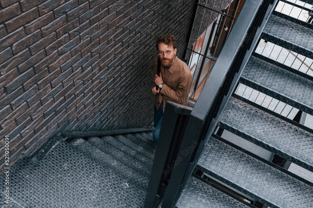 Looking up. Man in khaki colored jacket and in jeans is on the stairs on the building outdoors