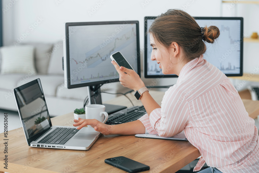 Using smartphone. Female stock broker in casual clothes is working in the office by pc Using smartphone. Female stock broker in casual clothes is working in the office by pc
