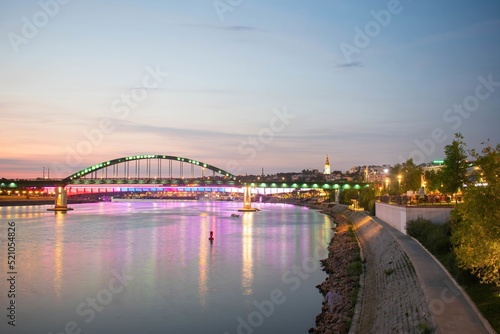 Belgrade Old Sava Bridge just after sunset lighted with LED photographed from river bank just below Belgrade Promenade in summer evening with Ortodox Church, Sava port and old town behind in the scene