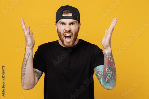 Young sad angry indignant furious bearded tattooed man 20s he wears casual black t-shirt cap spread hands scream shout isolated on plain yellow wall background studio portrait. Tattoo translate fun.