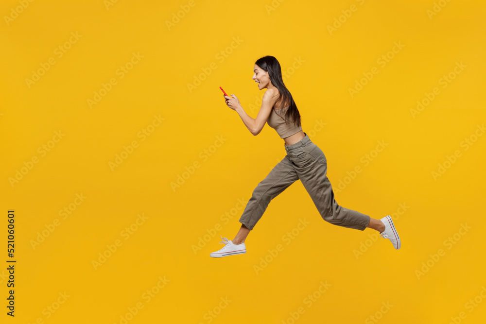 Full body side profile view young latin woman 30s she wear basic beige tank shirt jump high run fast use mobile cell phone isolated on plain yellow backround studio portrait. People lifestyle concept.