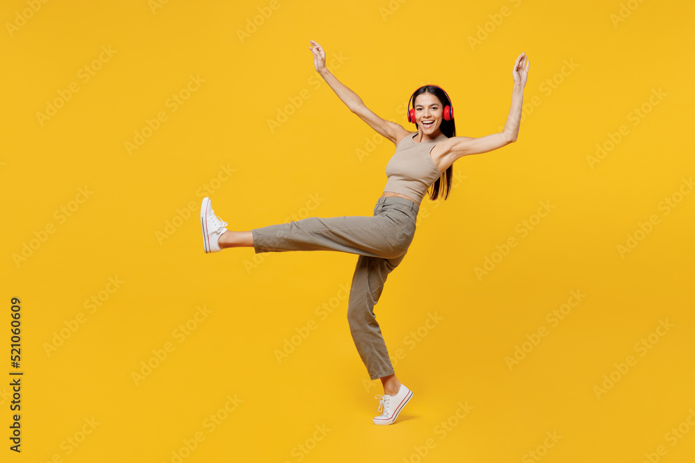 Full body happy young latin woman 30s she wear basic beige tank shirt headphones listen to music dance raise up hands leg isolated on plain yellow backround studio portrait. People lifestyle concept.