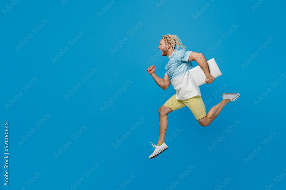 Full body young blond man with dreadlocks 20s he wear white t-shirt hold closed laptop pc computer jump high run fast isolated on plain pastel light blue background studio. People lifestyle concept.