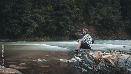 A man with a beard in a plaid shirt and shorts sits on the bank of a mountain river, moody colors