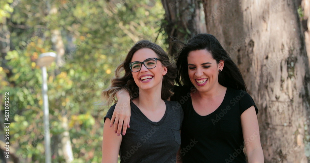 Two girlfriends together walking at the park embracing each other laughing Two girlfriends together walking at the park embracing each other laughing