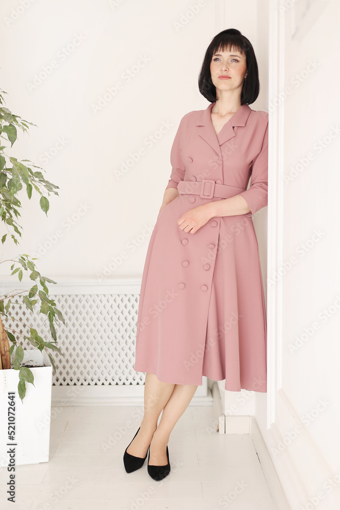 Portrait of attractive smiling brunette with short hair woman staying alone on white stylish wall background with green houseplant. Female in soft pink dress is looking in camera Portrait of attractive smiling brunette with short hair woman staying alone on white stylish wall background with green houseplant. Female in soft pink dress is looking in camera