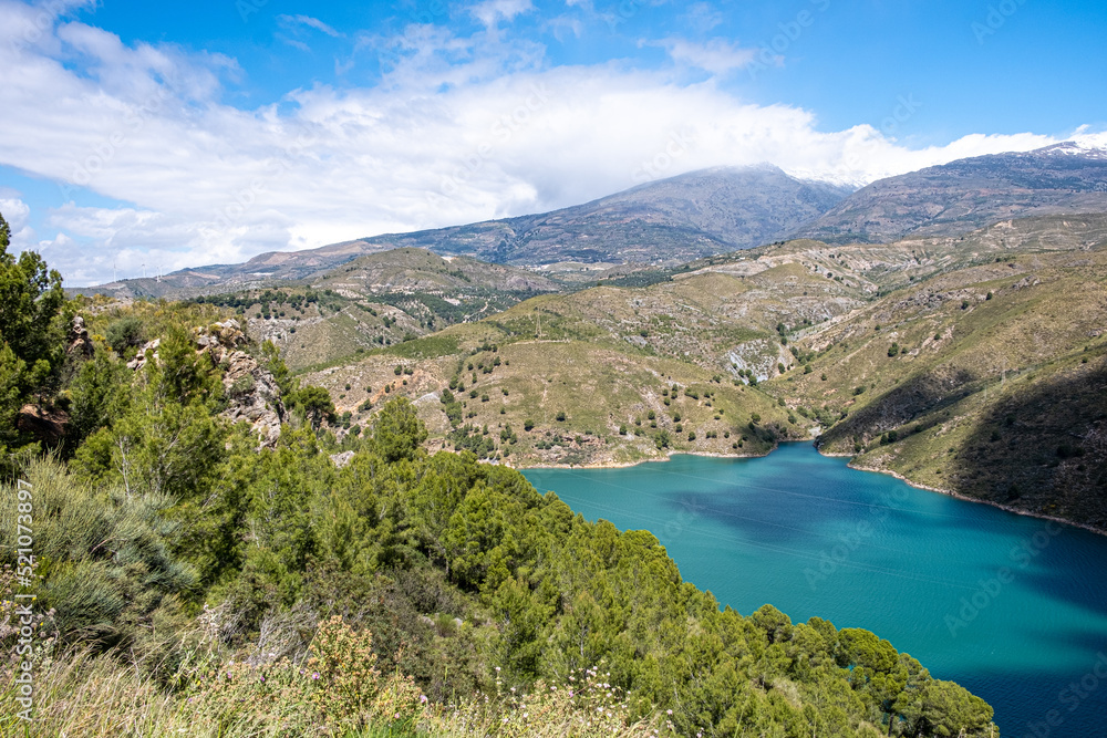 View from the top on green lake surrounded by picturesque mountain alley with trees and bushes with blue sky and white clouds in background in sunny summer day. View from the top on green lake surrounded by picturesque mountain alley with trees and bushes with blue sky and white clouds in background in sunny summer day.