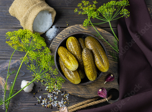 Pickled cucumbers in a clay bowl on a wooden plank. Salt in a canvas bag and sprigs of dill in the background. Dark brown napkin on the table. Top view