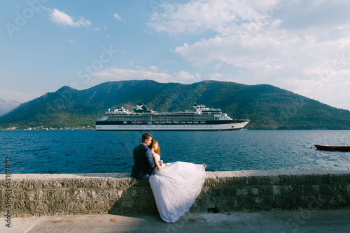 The bride and groom are sitting hugging on a stone fence of the pier, the bride is leaning on the groom, cruise liner behind them
