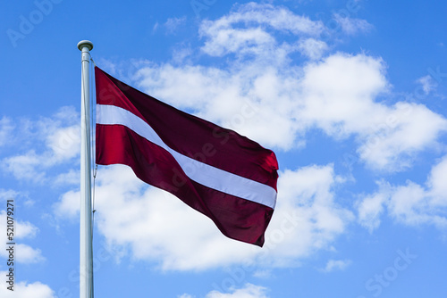 Latvian National Flag on the mast waving in the wind against blue sky with white clouds