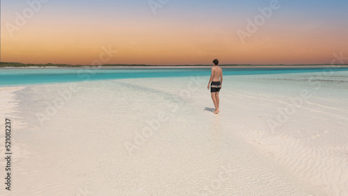 Young man standing from behind on tropical white sand beach in Caribbean looking over the perfect turquoise ocean. Luxury living vacation destination.