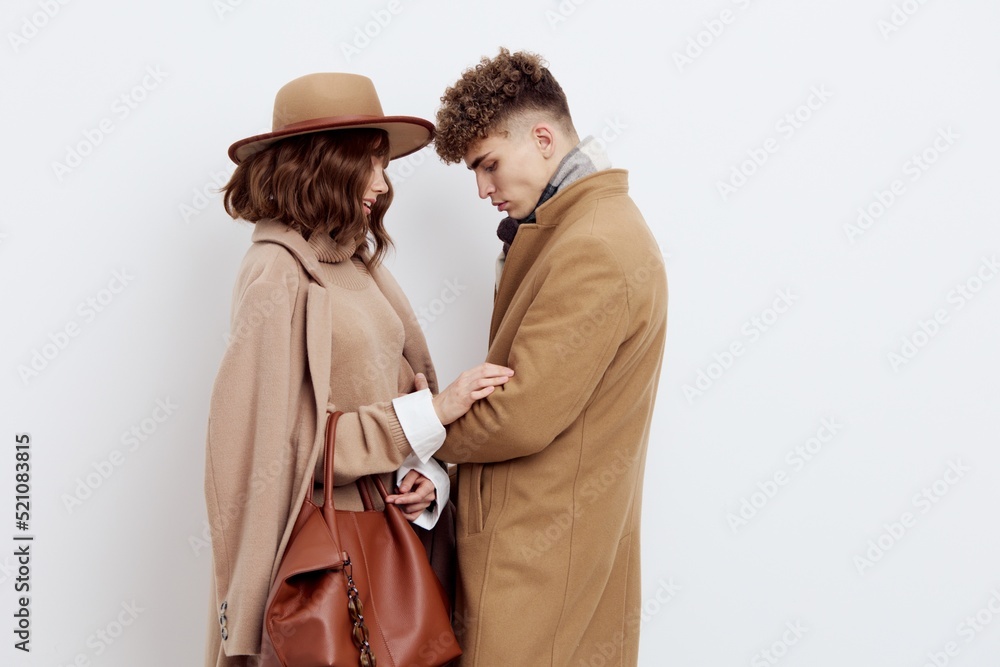 a cute couple stands on a light background dressed in coats and sweaters, a man stands against a woman and hugs her in front a cute couple stands on a light background dressed in coats and sweaters, a man stands against a woman and hugs her in front