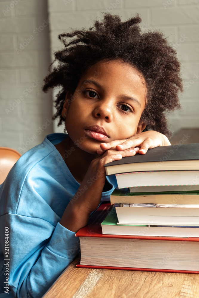 Sad tired African-American girl doing homework at home,puting her head on a stack of textbooks.Back to school concept.School distance education,home schooling,diverse people. Sad tired African-American girl doing homework at home,puting her head on a stack of textbooks.Back to school concept.School distance education,home schooling,diverse people.