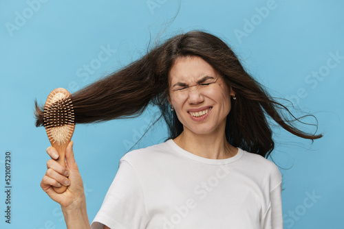 middle-aged brunette woman combing her long beautiful hair with a wooden comb and screaming loudly in pain while standing on a blue background with empty space for text
