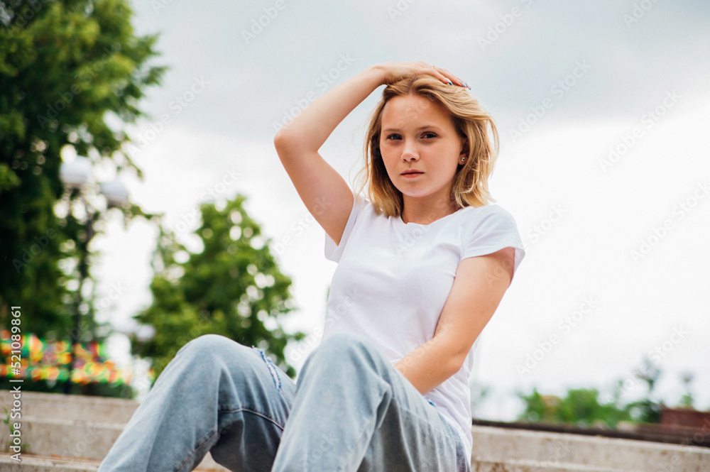 Portrait of a teenage girl in casual clothes in the park. Summer skateboarding, active lifestyle. A student or a schoolboy is sitting on a skateboard during the summer holidays. Sports recreation.