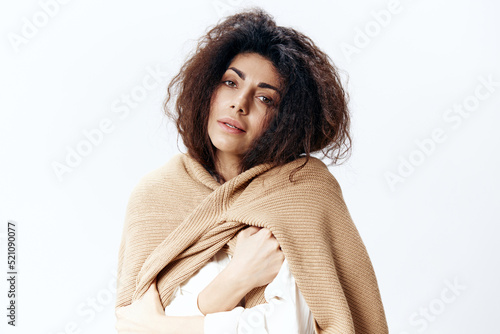 Serious Curly Pretty Latin Woman in Beige Warm Sweater White Shirt Hugging Herself Posing Isolated Looking At Camera At White Studio Wall Background. Fashion Seasonal Sale New Collection Concept