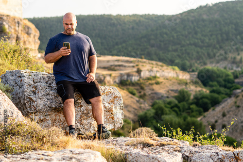 Foto Stocky man using a phone leaning on the rock of a mountain.