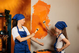 Woman with little girl using orange paint on walls and brush to renovate house interior, painting apartment room together. People having fun with housework redecoration, diy decorating job.