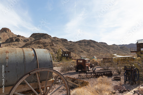 1932 American sedan sits rusting near wagon and vintage school bus under the hot desert sun among the mining town wasteland turned hidden ghost town