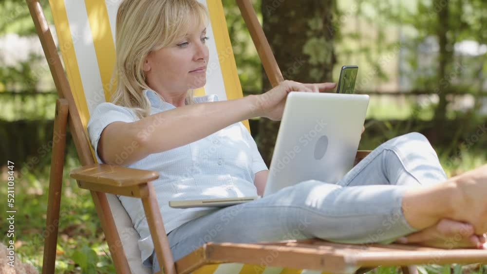 Relaxed woman uses computer laptop and mobile phone for surfing internet to smart working or shopping online. Outdoor in green home garden in sunny summer day sitting on the deck chair