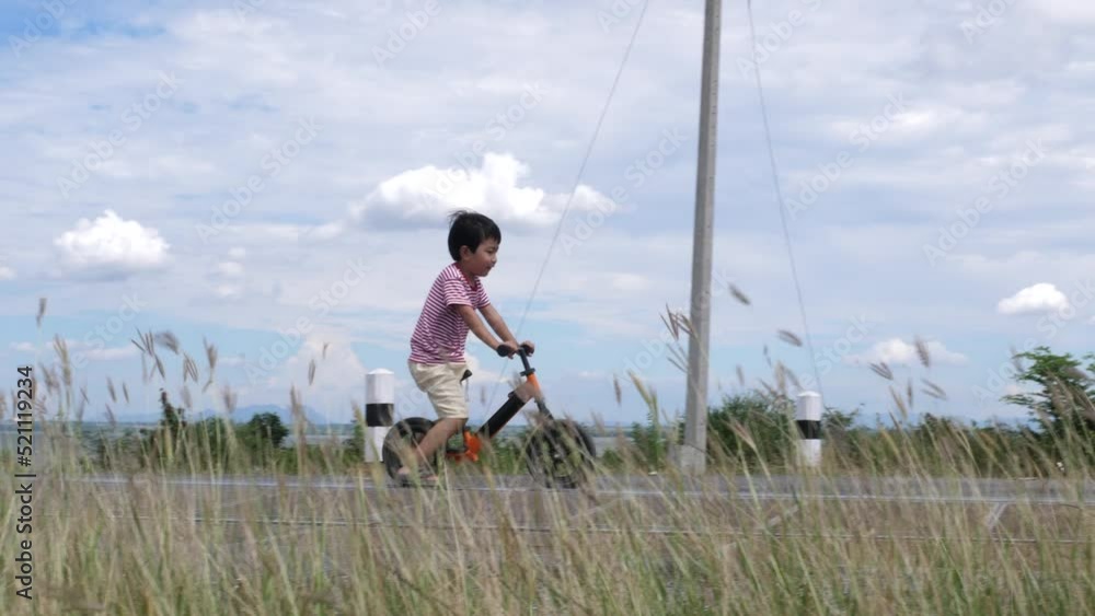 Asian child boy riding bicycle on road outdoors in blue sky background, grass swaying by wind. Kid learning to cycling with fun. Concept of happiness, freedom, sport, family activity, health care. Asian child boy riding bicycle on road outdoors in blue sky background, grass swaying by wind. Kid learning to cycling with fun. Concept of happiness, freedom, sport, family activity, health care.