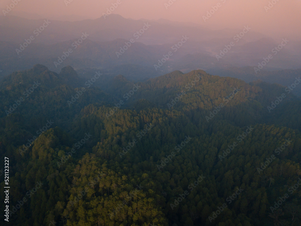 A panoramic view of the foggy Mountains with dense of forest. Orange sky with clouds over layers of green hills and mountains. Copy space.  Menoreh Hill, Central Java, Indonesia