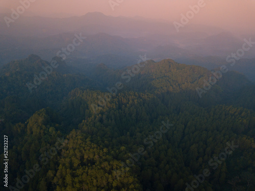 A panoramic view of the foggy Mountains with dense of forest. Orange sky with clouds over layers of green hills and mountains. Copy space. Menoreh Hill, Central Java, Indonesia