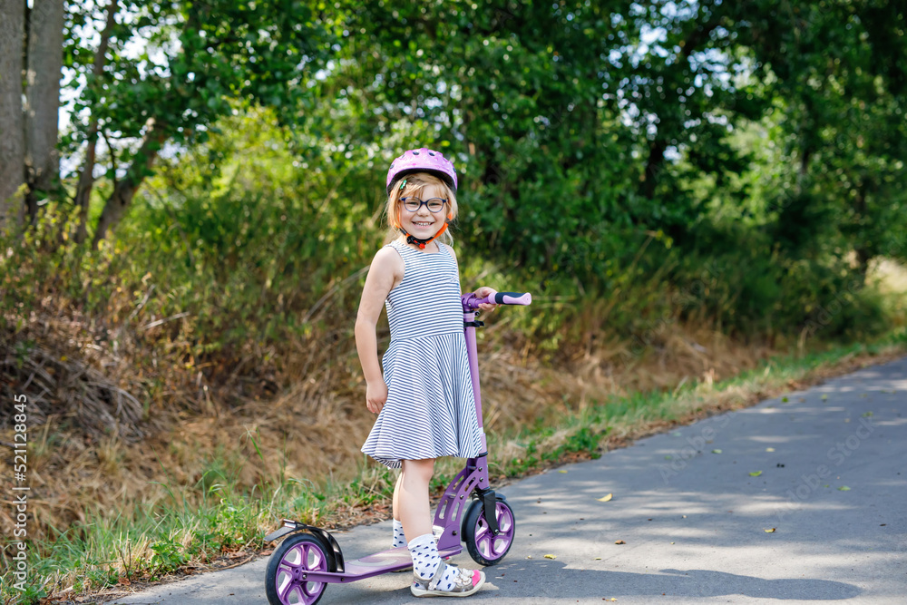 Fototapeta premium Portrait of active little preschool girl with glasses and helmet riding scooter on road in park outdoors on summer day. Seasonal child activity sport. Healthy childhood lifestyle