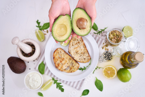 Avocado and cream cheese toasts preparation - Woman holding halved avocado over table with ingredients