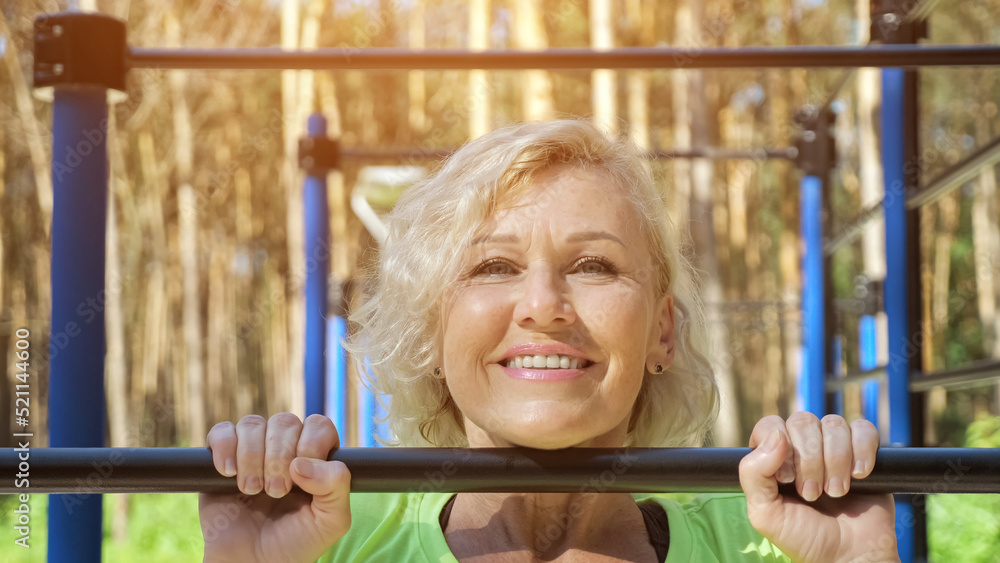 Obraz premium Mature woman does pull-ups smiling on park sports-ground in sunny morning. Young blonde pensioner wearing yellow top enjoys healthy lifestyle, closeup