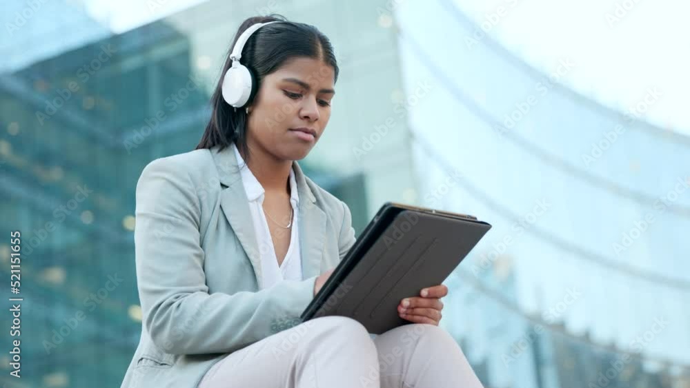 Serious business woman typing on a tablet and listening to music using headphones while sitting outside a building. Female corporate professional browsing the internet or writing an email outdoors Serious business woman typing on a tablet and listening to music using headphones while sitting outside a building. Female corporate professional browsing the internet or writing an email outdoors