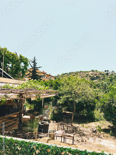 Old abandoned mediterranean farm house surrounded by trees. Sun lit shabby farmhouse with old fashioned wooden furniture chairs in the garden.