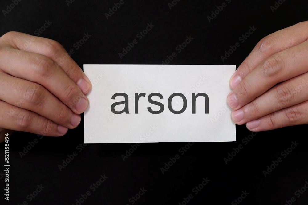 Arson crime case mug shot concept. Criminal hands holding paper placard with written word in dark black background. Arson crime case mug shot concept. Criminal hands holding paper placard with written word in dark black background.