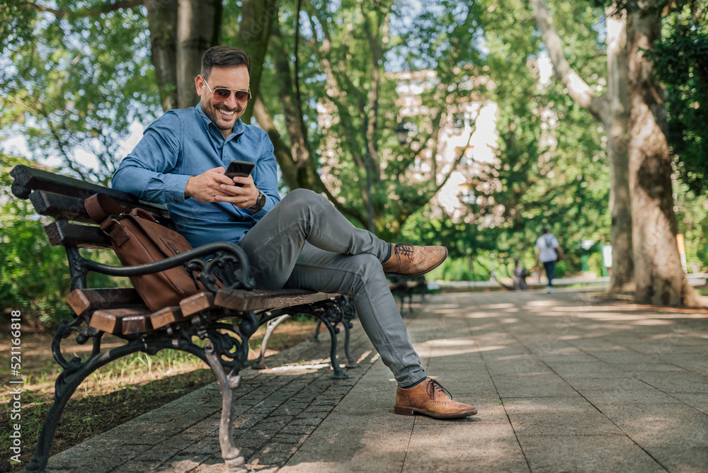 Fototapeta premium Smiling businessman networking on smart phone while sitting on bench at the park