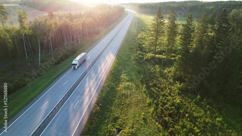 White semi-trailer truck delivers a load, driving down a scenic country highway through beautiful morning woods in the dawn light. Delivery, Logistics, Freight, Trucking Concept. Aerial view. photo