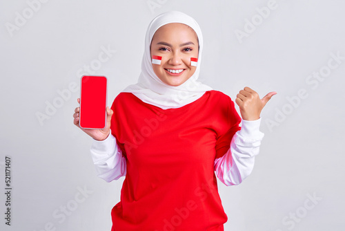 Cheerful young Asian muslim woman in red white t-shirt showing blank screen mobile phone and pointing aside isolated on white background. celebrating indonesian independence day on 17 august