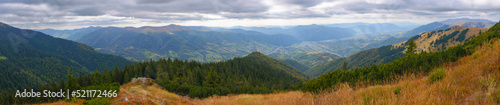 panorama of mountainous countryside in autumn. colorful grassy meadows and spruce forest on the hillside. view in to the rural valley in dappled light. ridge beneath a cloudy sky in the distance