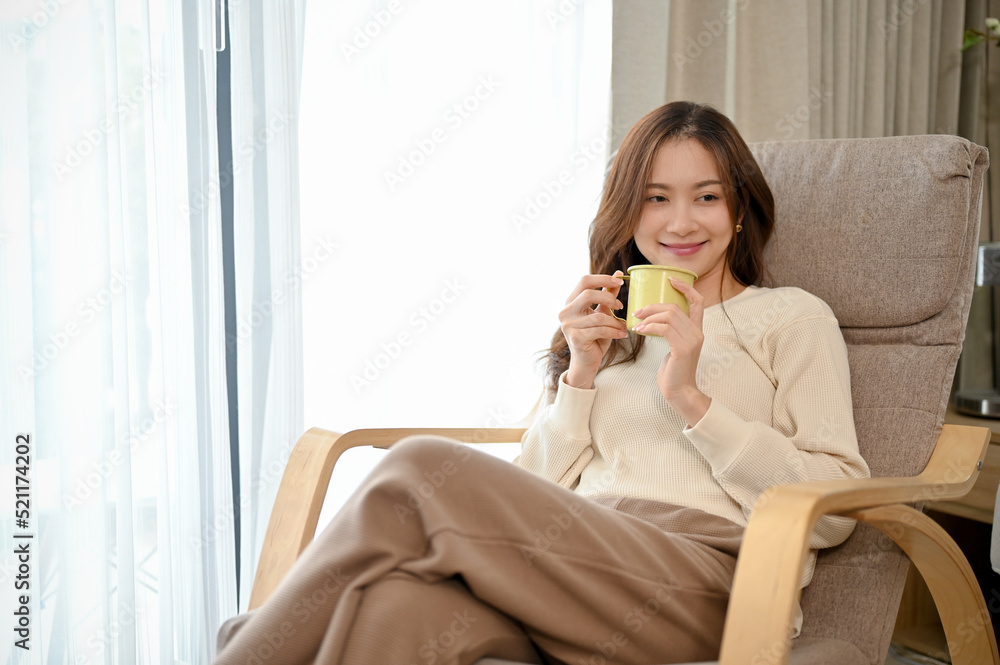 Young Asian female relaxes in living room, sits on comfy armchair and sips hot coffee. Young Asian female relaxes in living room, sits on comfy armchair and sips hot coffee.