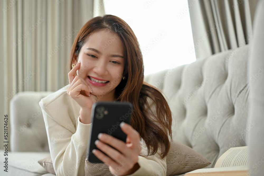 Attractive Asian female laying on sofa and chatting with her friends on her smartphone. Attractive Asian female laying on sofa and chatting with her friends on her smartphone.