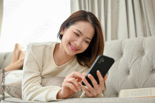 Charming young Asian female laying on her comfy sofa, chatting with her friends on smartphone.