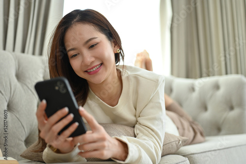 Relaxed Asian female laying on comfy sofa in the living room, using smartphone