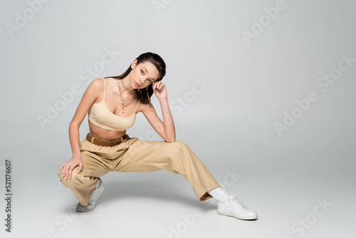 full length of brunette woman in stylish beige outfit and trendy accessories posing on grey