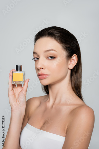 pretty woman with bare shoulders holding bottle with luxury perfume isolated on grey