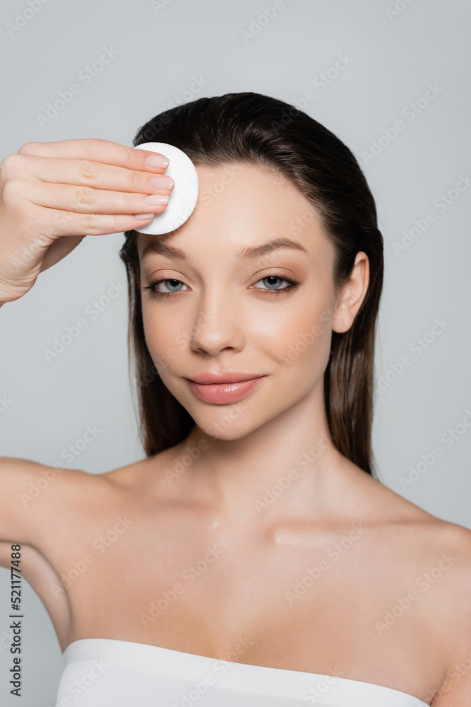 happy young woman removing makeup on forehead with cotton pad isolated on grey happy young woman removing makeup on forehead with cotton pad isolated on grey