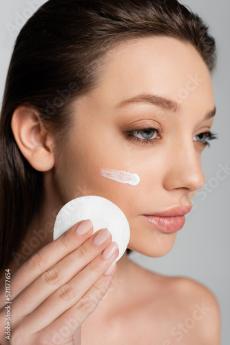 close up of young woman with cosmetic product on face holding cotton pad isolated on grey