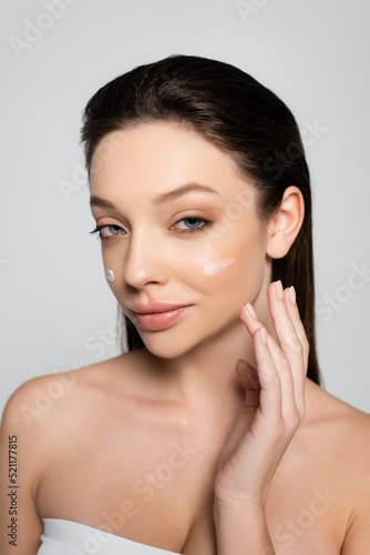 young woman with bare shoulders and cream on cheeks smiling isolated on grey