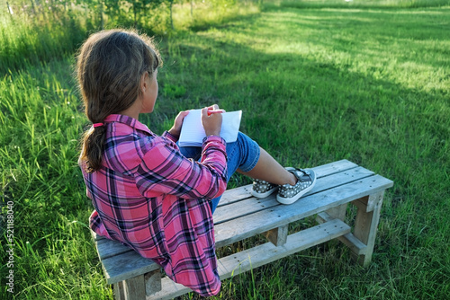 Young girl sitting on bench and writing on notebook outdoor, abstract blurred sunny natural background. Child studying in garden. education, writing hobby concept
