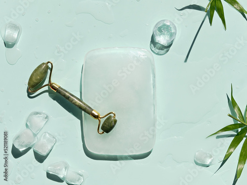 Jade stone face roller and self made moisturizer with ice cubes and exotic palm fern leaves on mint green background. Facial massage concept, handmade cosmetics.