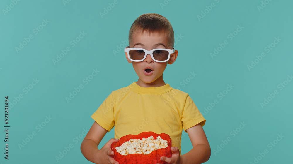 Obraz premium Little toddler children boy in t-shirt eating popcorn and watching comedy movie film wearing 3D glasses, enjoying cinema snacks. Young preschool kid in glasses isolated alone on blue studio background