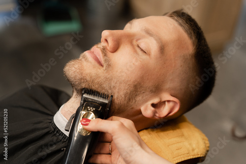 a man smiles while trimming his beard with a hair clipper. light unshavenness.